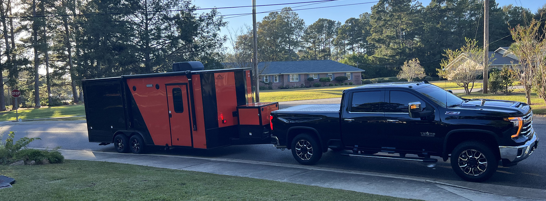 The image shows two trucks parked side by side on a street, with one being a large black truck and the other a smaller red truck pulling a trailer with a black roof.