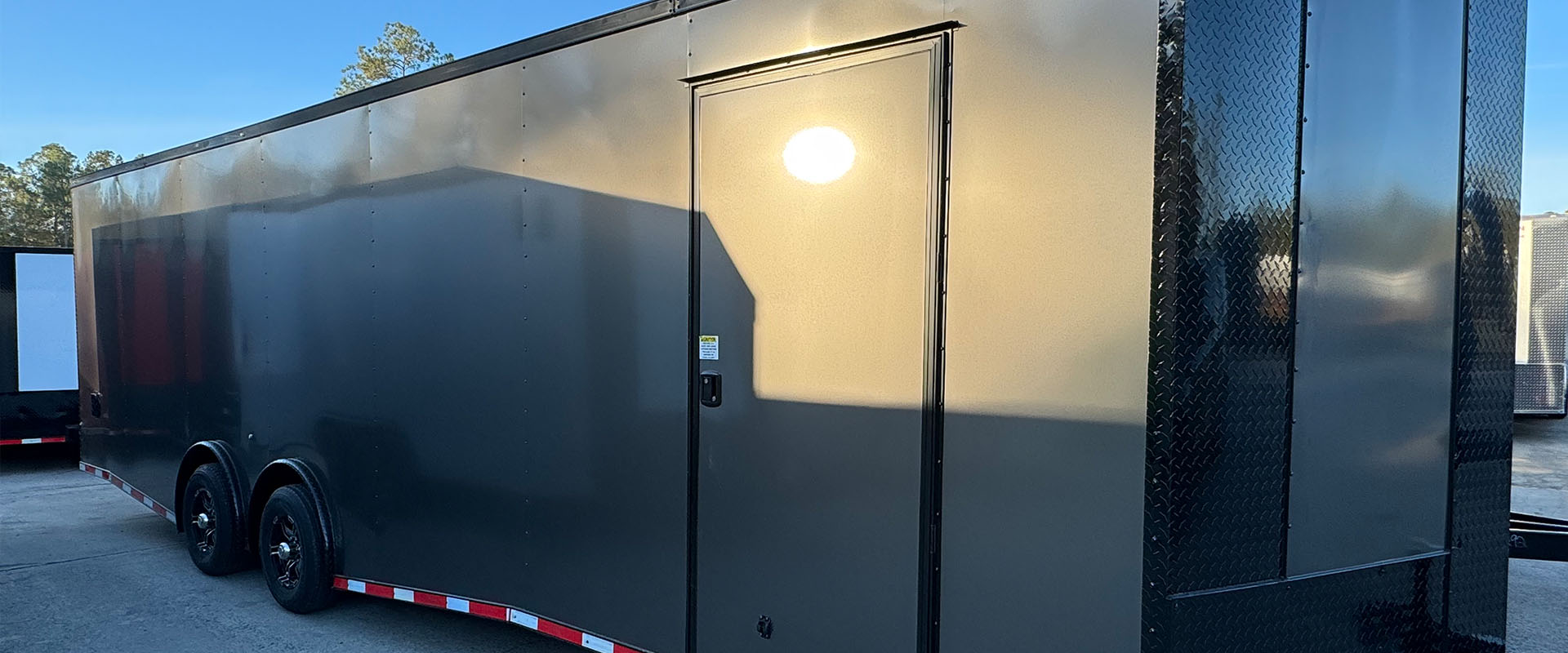 The image shows a large black trailer parked on a concrete surface with a clear sky background.