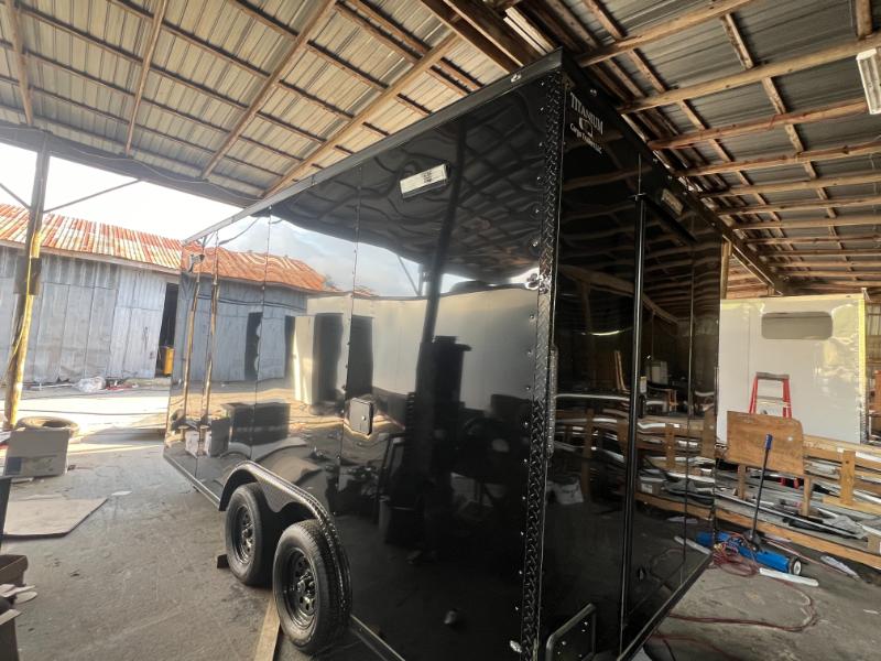 The image shows a large black trailer parked inside a building with a metal roof, next to a wall with corrugated metal panels.