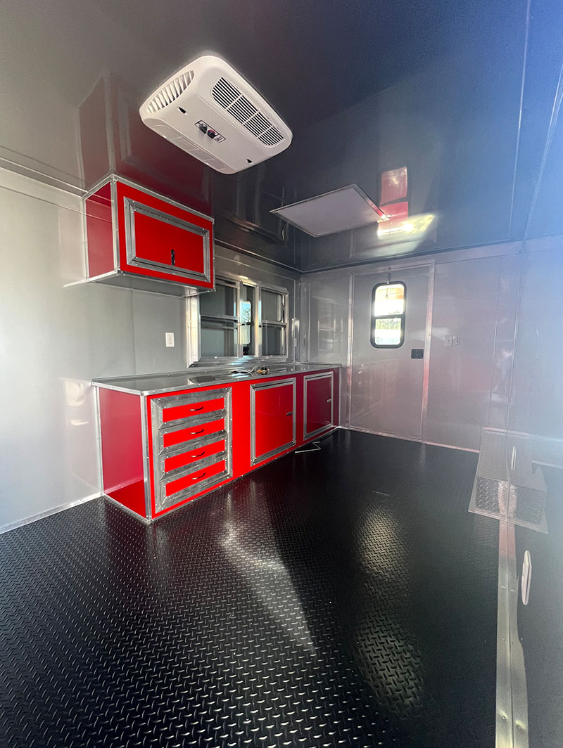 The image shows the interior of a large commercial kitchen with red and white cabinets, black flooring, stainless steel appliances, and a central island with a sink.