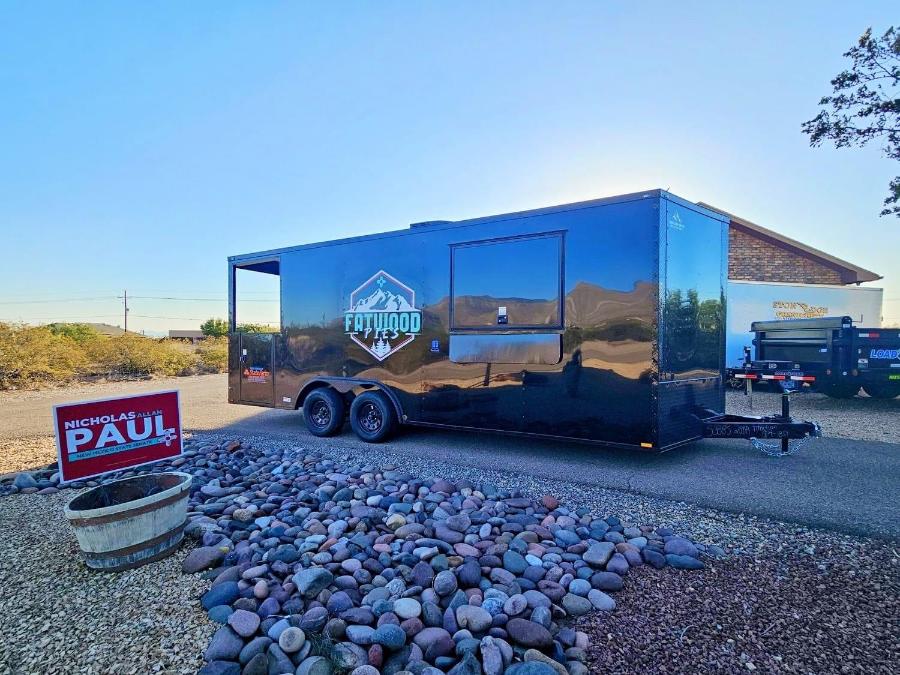 The image shows a black enclosed trailer parked on a gravel surface with a sign nearby, under a clear sky.