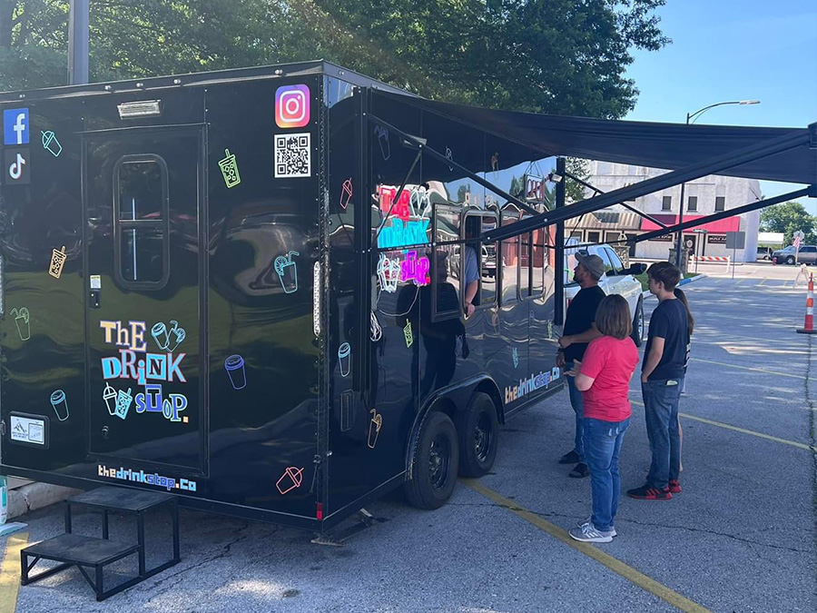 The image shows a large black food truck parked outdoors during daylight with people standing nearby, featuring a colorful sign on its side indicating The Drink Up and various decorative elements, including stickers and graffiti-like designs.