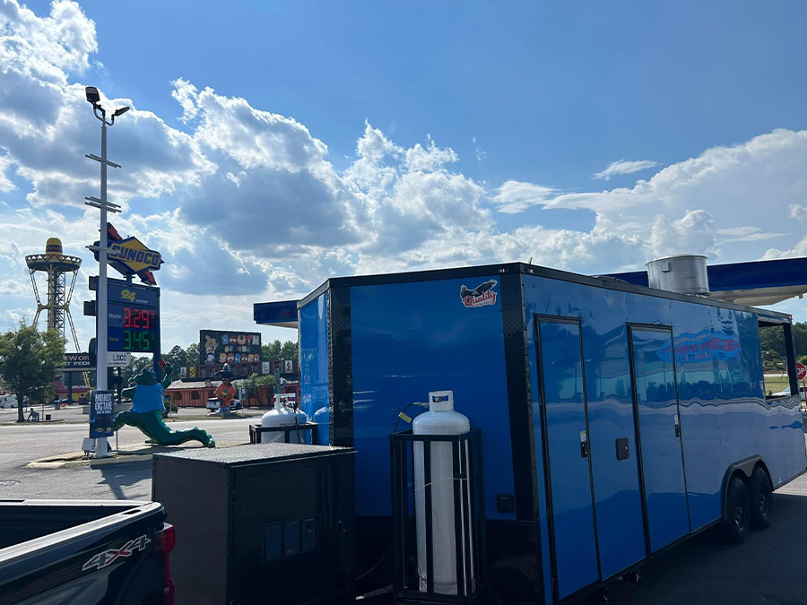 The image shows a large blue food truck parked next to a gas station with its side door open, under a sunny sky.