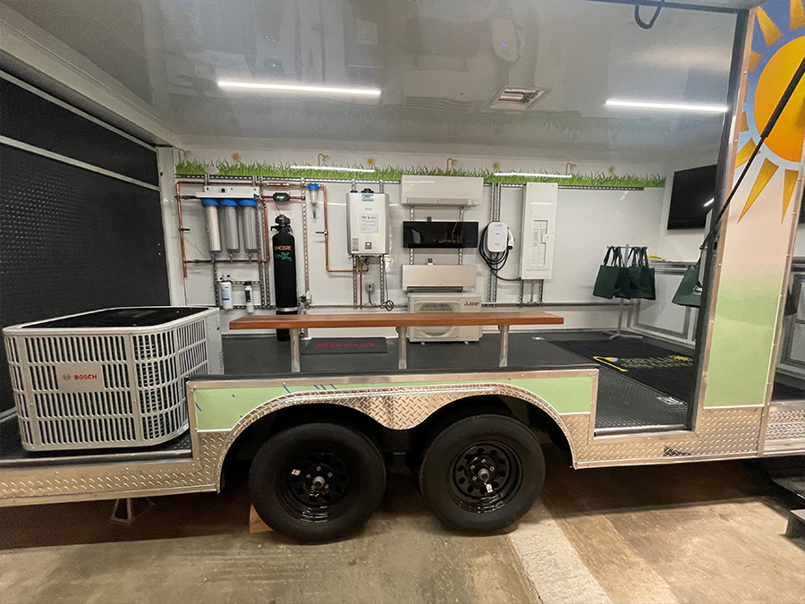 An image of a large, green and white trailer parked indoors with its doors open, showcasing an interior filled with various equipment and appliances, including a sink and oven.