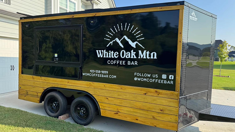 The image shows a mobile food truck, painted in black and yellow with a logo for White Oak Mtn and an illustration of mountains on its side. It s parked outside a house under clear skies.