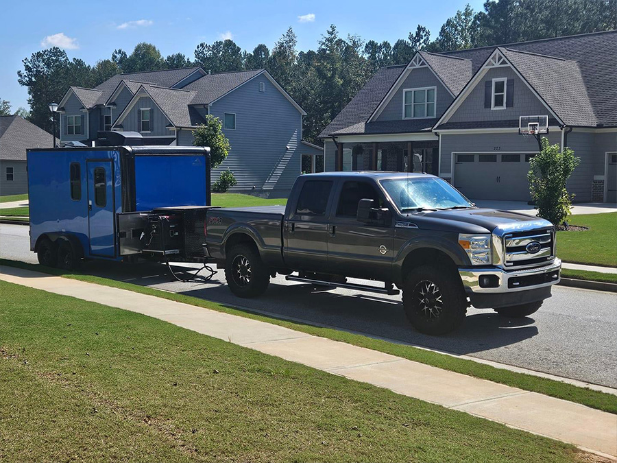 A large blue truck with a camper shell attached, parked on a residential street next to a house.