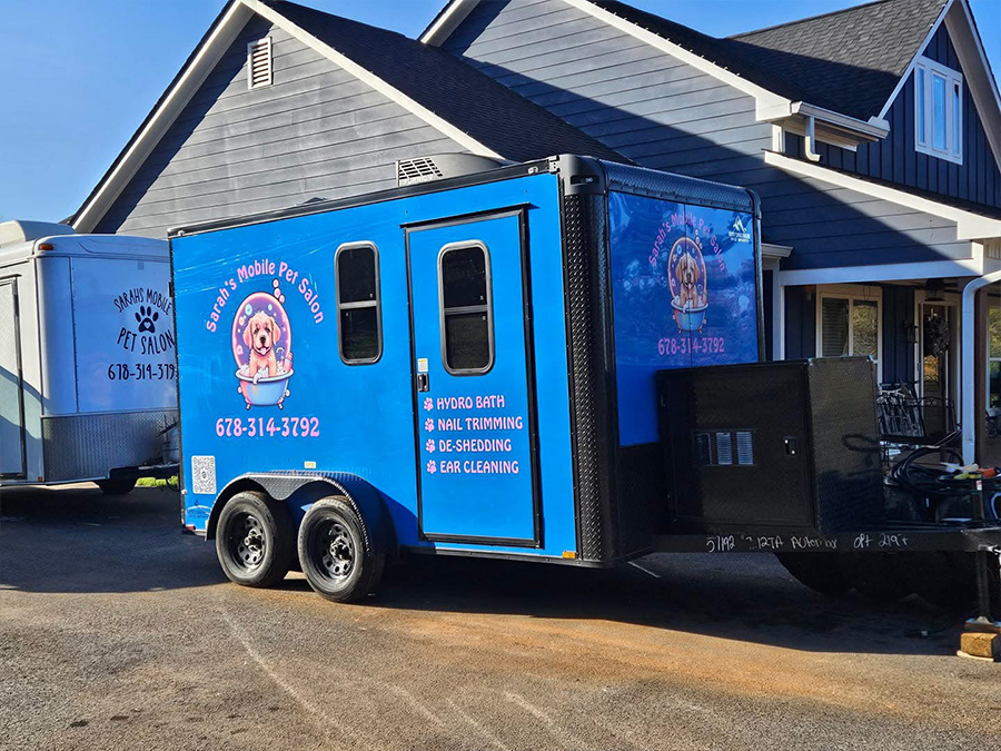 This is an image of a blue trailer parked outside a building with a sign that reads Mobile Pet Grooming. The trailer has a logo on its side, which appears to be related to pet grooming services.