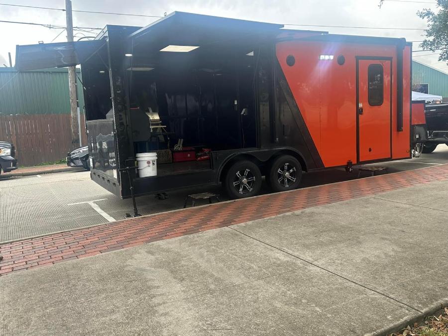 The image shows a large, red and black mobile catering truck parked on a street with a brick sidewalk and a vehicle behind it.