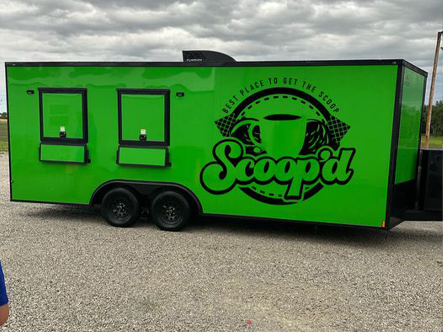 The image shows a large green food truck with a white logo featuring a coffee cup and the word 'Scoopit' on its side, parked on a gravel surface.