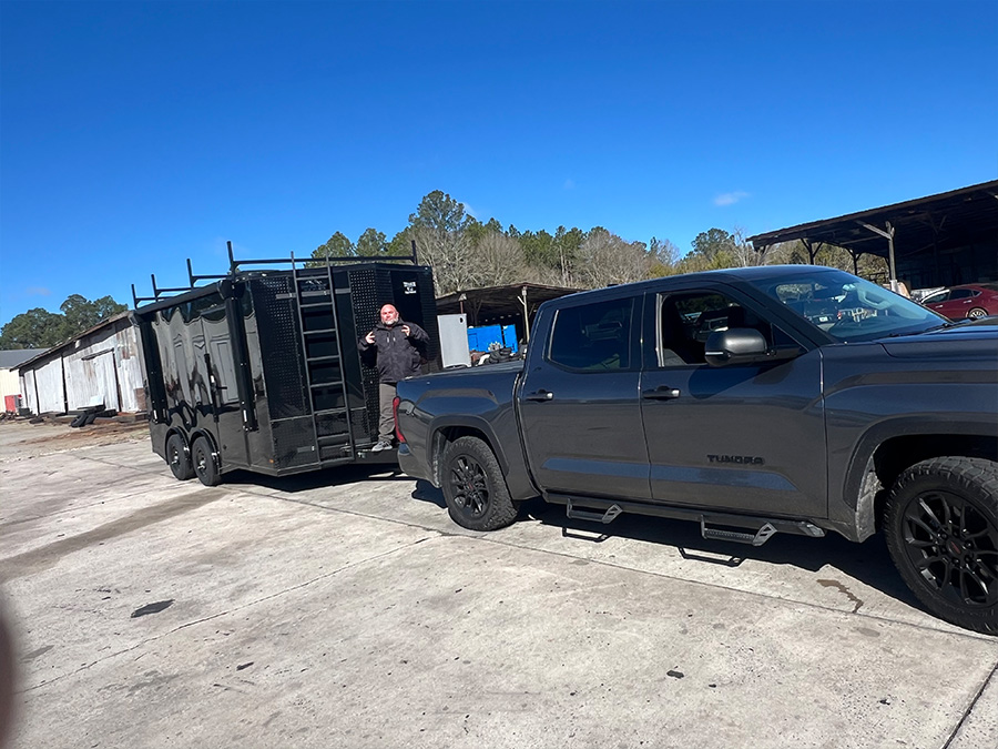The image shows a black pickup truck parked next to a trailer with a large black metal structure on top, possibly for transporting equipment or materials.