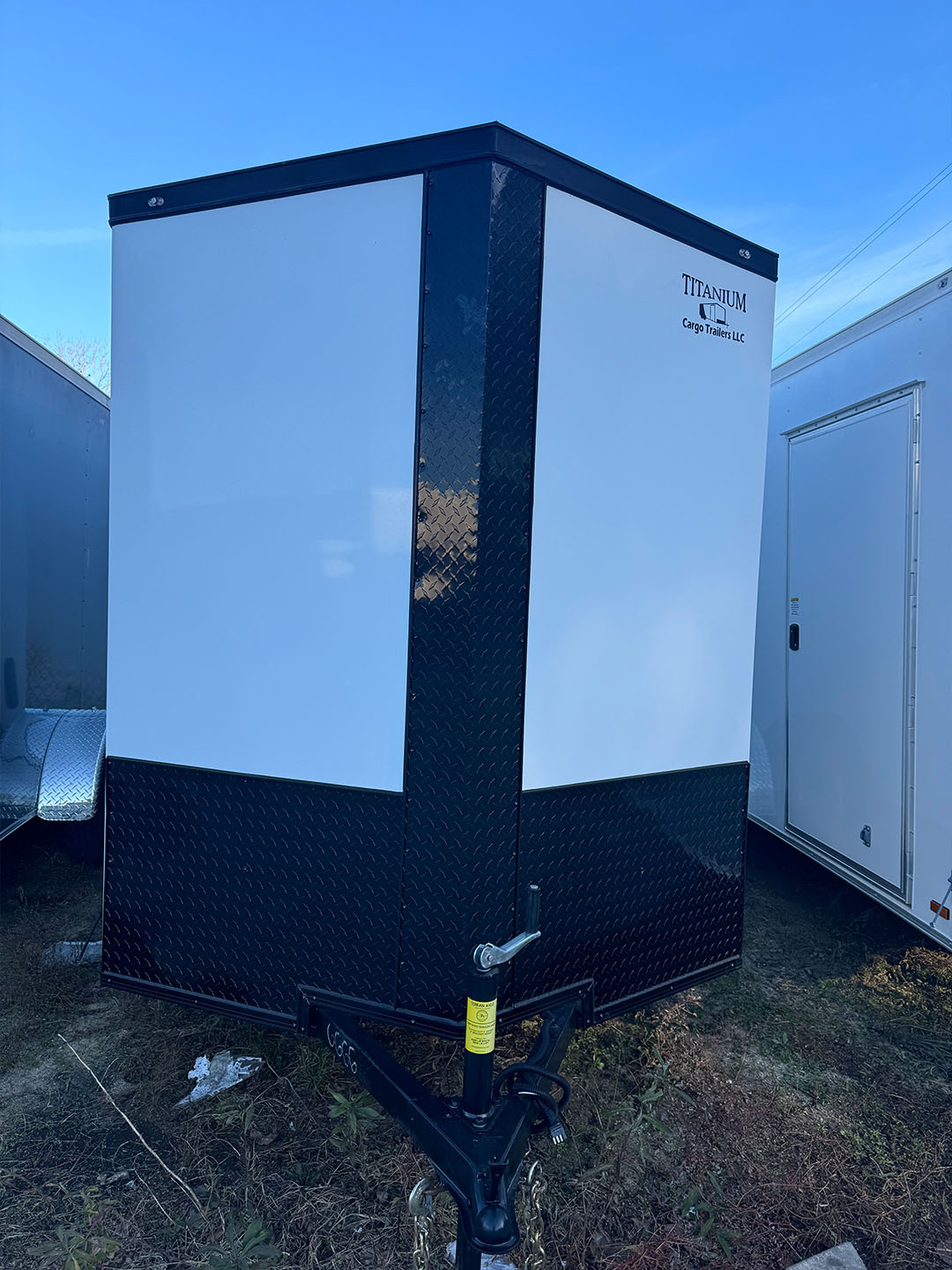 The image shows a portable toilet with a black exterior and a white door, placed outdoors on a grassy area next to a trailer.