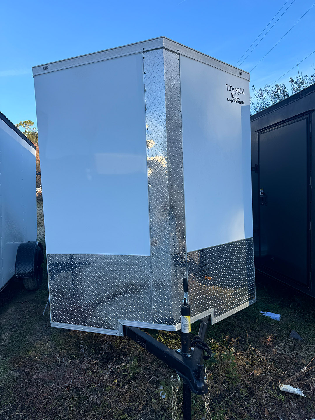 The image shows an outdoor portable restroom unit with a white exterior and a silver metal door, parked on a grassy area near a building with trailers.