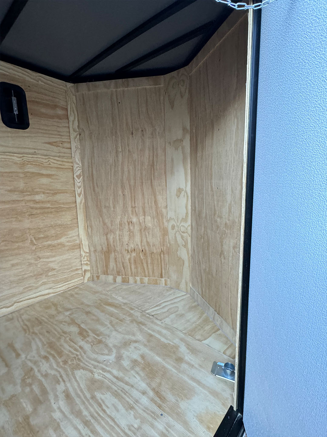 The image shows an interior view of a trailer with wooden walls and flooring, featuring a black plastic container at the end.