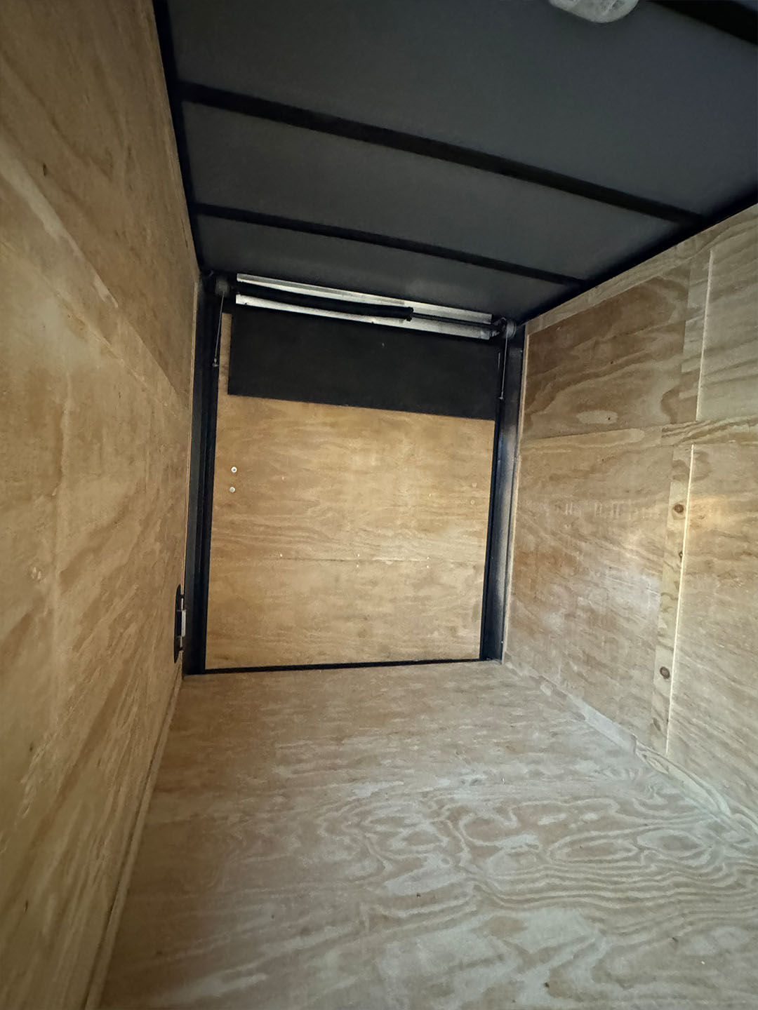 The image shows an interior view of a cargo trailer with cardboard walls, a wooden floor, and a black door at the front.