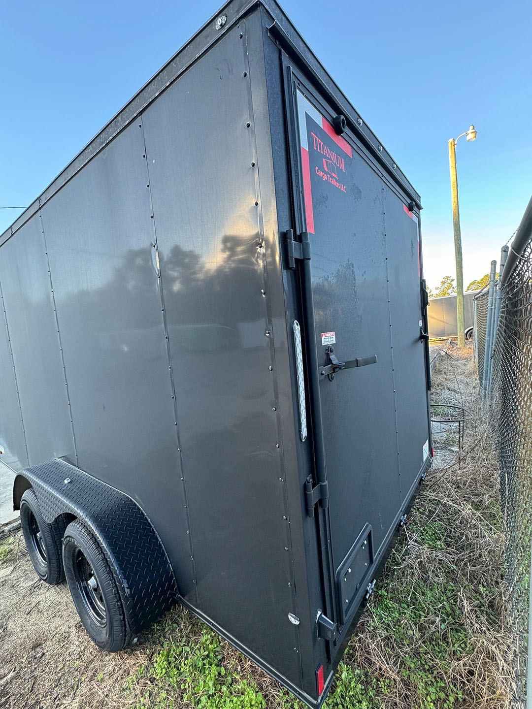 The image shows a large black cargo trailer parked on grass with its door open, under a clear blue sky.