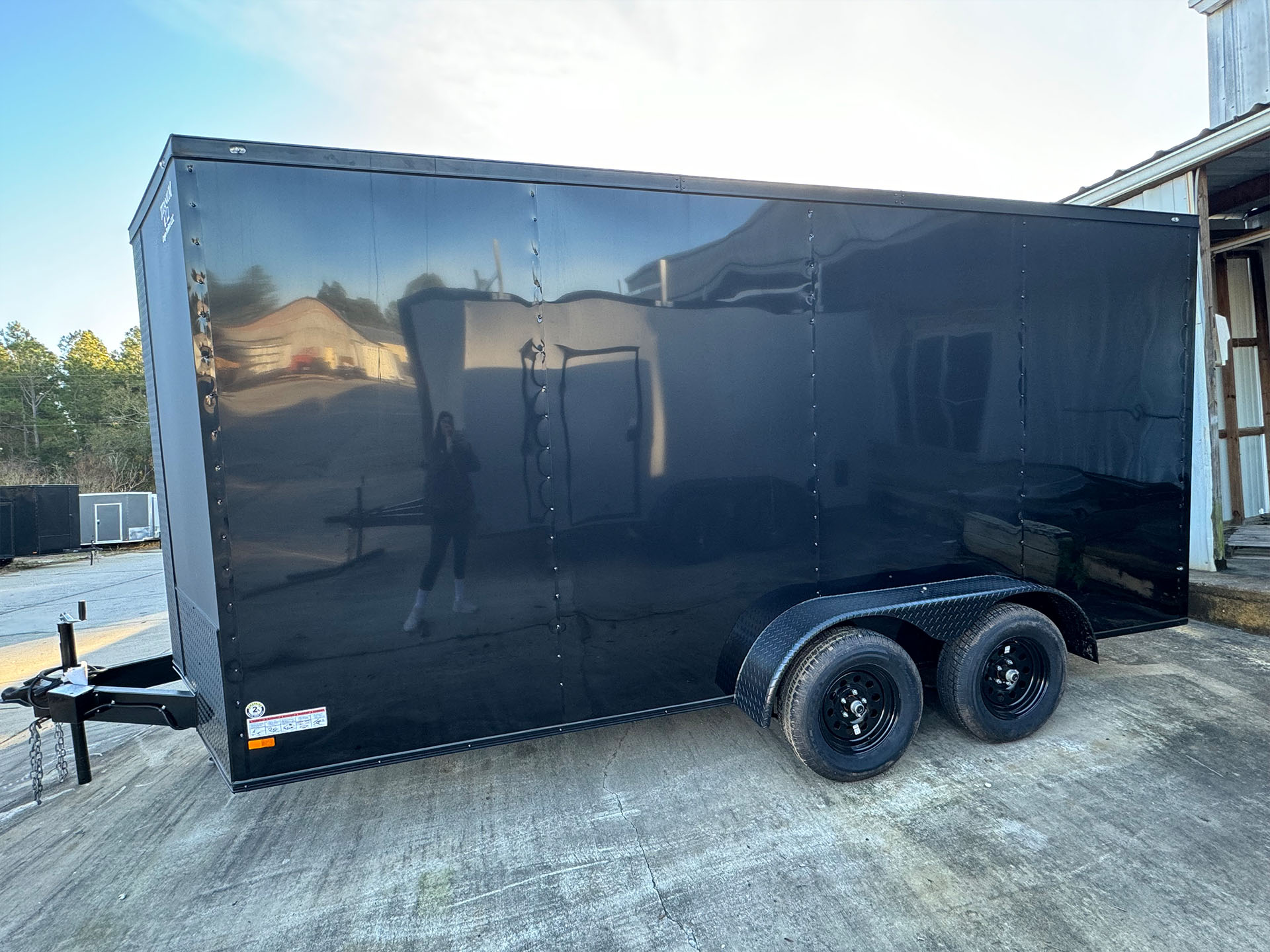 The image shows a black enclosed trailer parked on an asphalt surface with a person visible in the background.