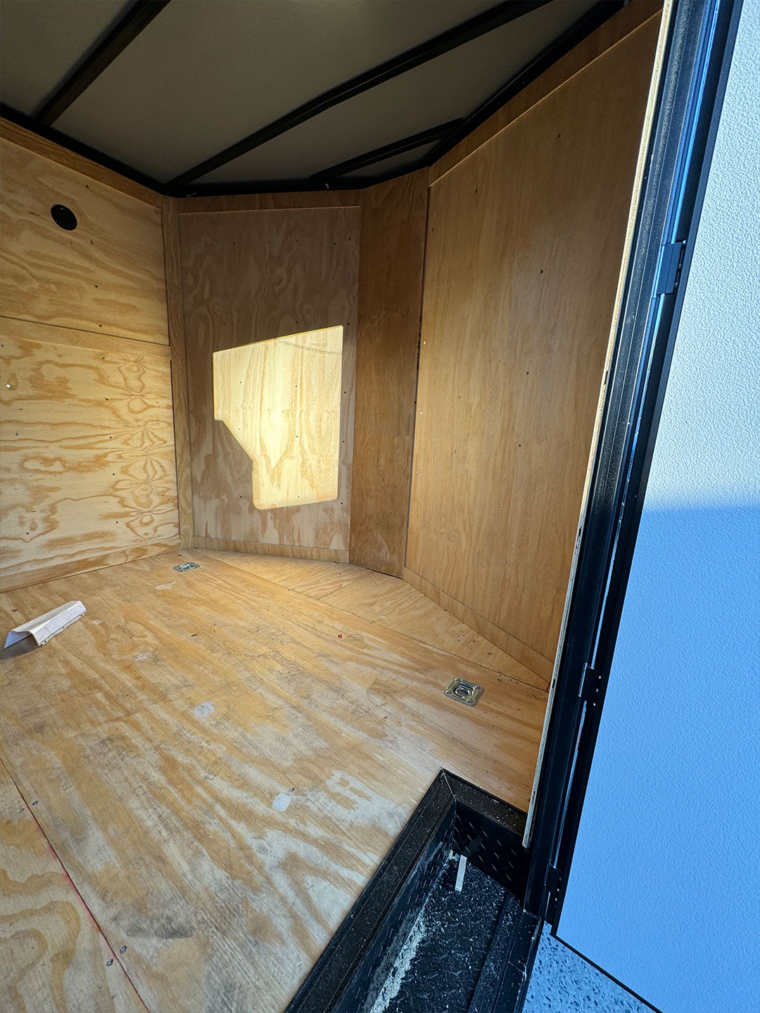 The image shows an empty interior of a trailer with a visible window, a simple door frame, and a cardboard box on the floor.