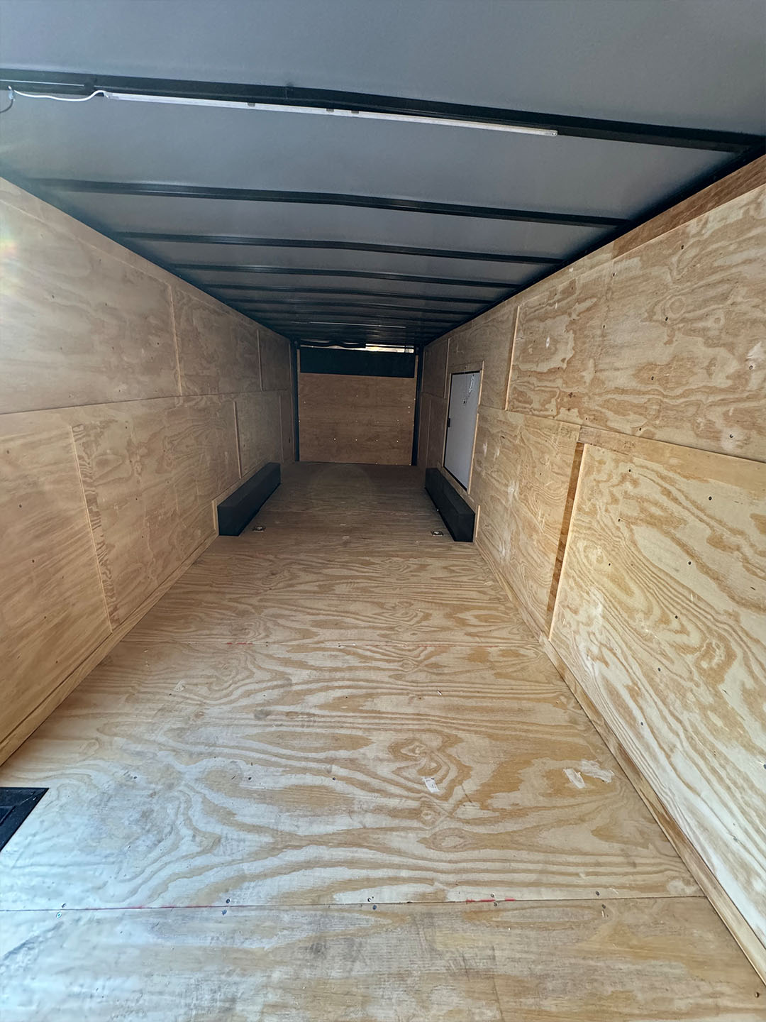 The image shows an empty shipping container with wooden flooring, viewed from the perspective of someone standing inside looking out towards a brightly lit area.