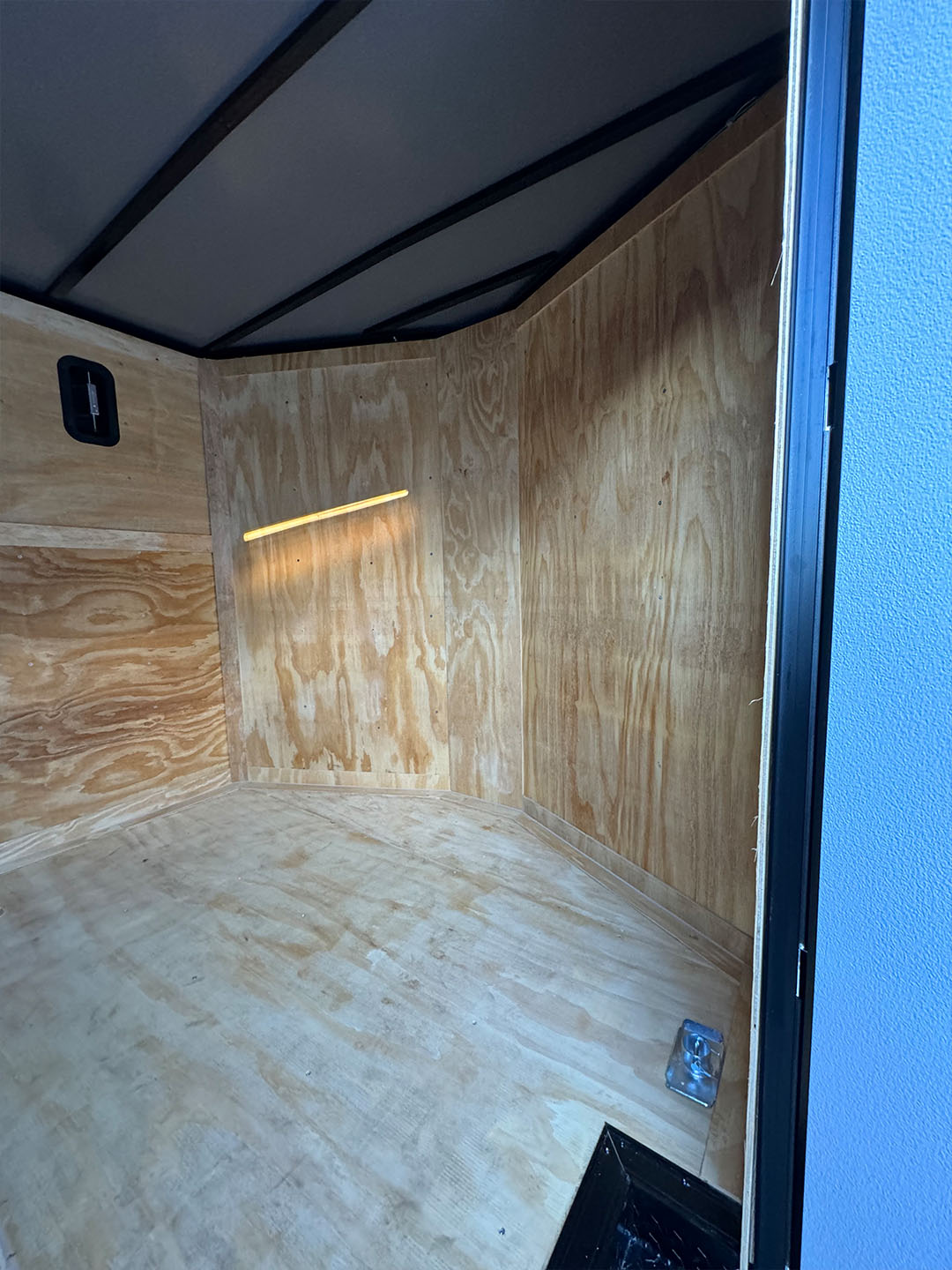 The image shows an empty, unfinished interior of a vehicle with plywood walls and floor, resembling a van conversion.