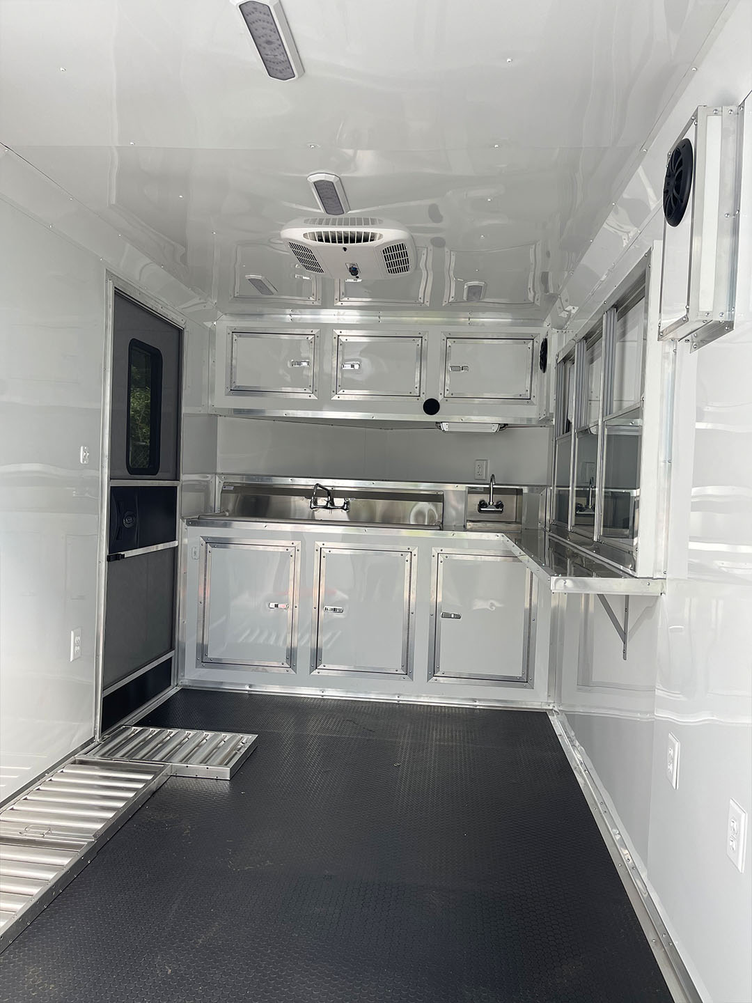 The image shows a large, empty, metallic food truck with stainless steel countertops and appliances, a sink, and an oven, parked inside a building with a black floor.