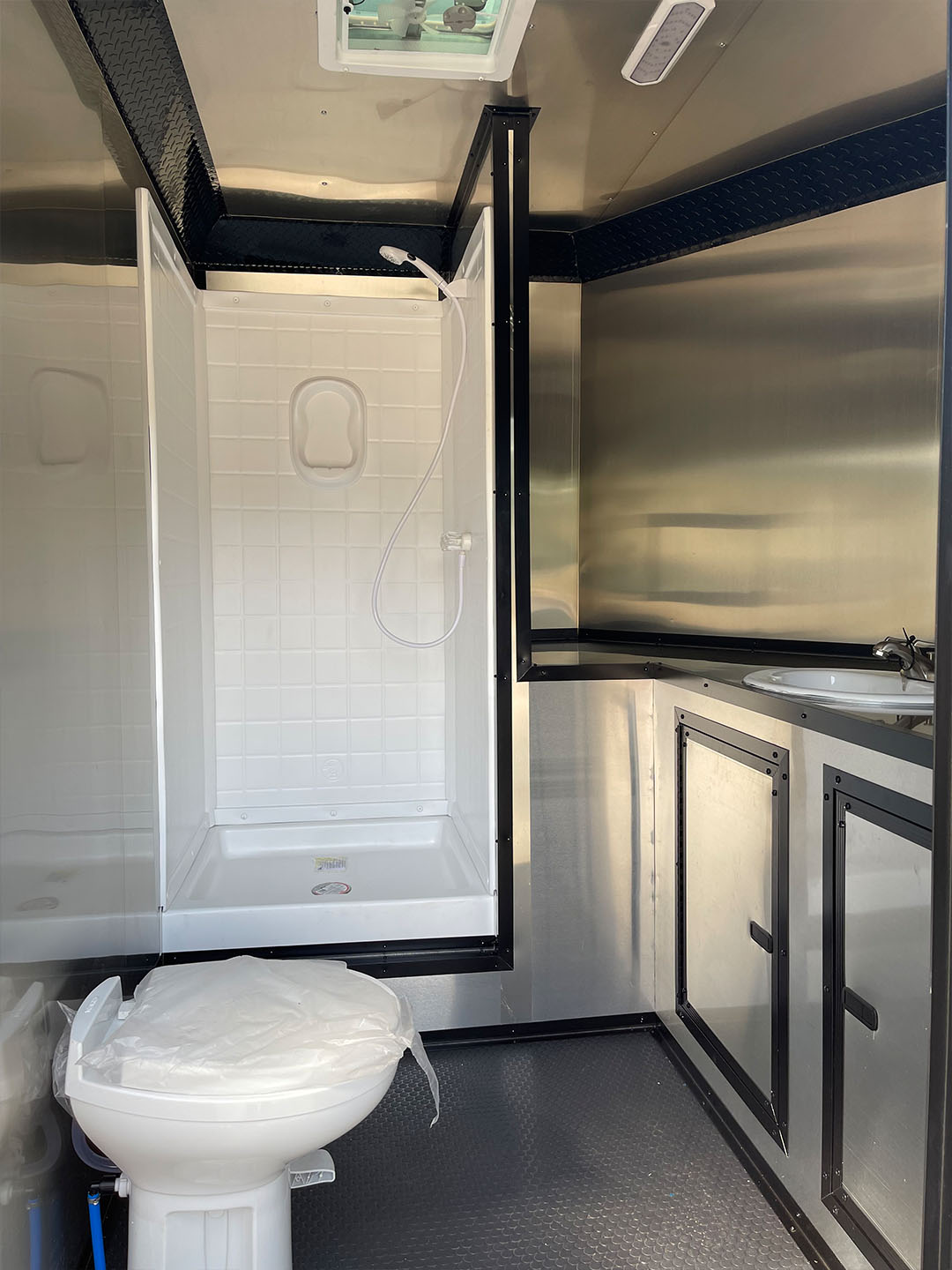 The image shows an interior view of a bathroom within a vehicle, featuring a shower stall with clear glass doors, a white toilet, and stainless steel fixtures.