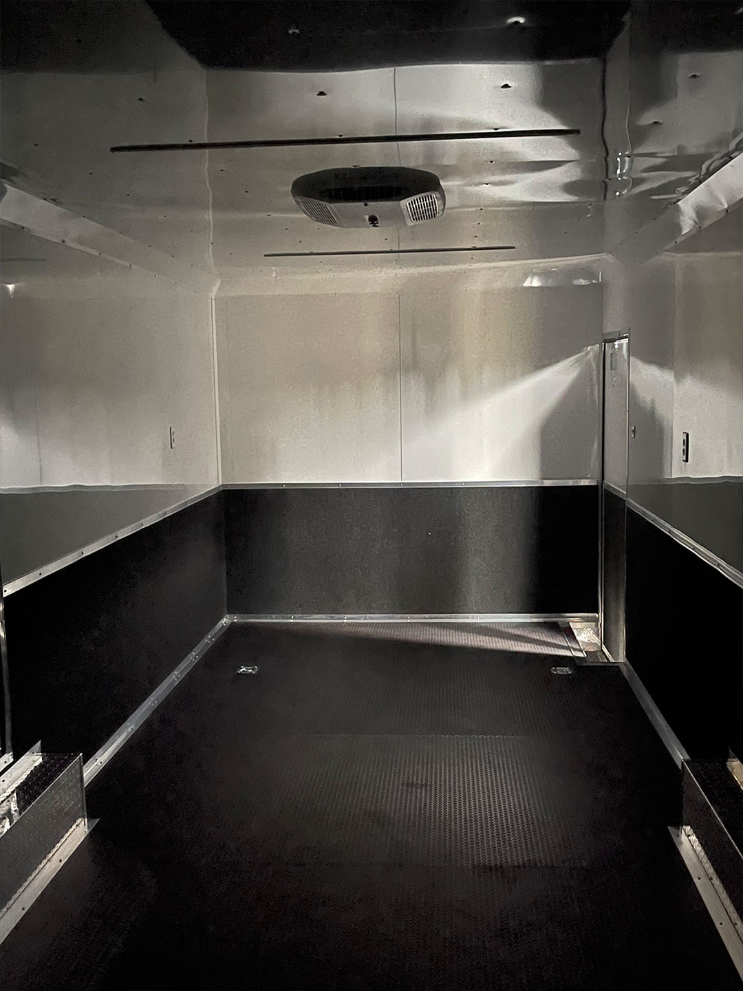 The image displays the interior of a trailer with metallic walls and a black floor, featuring a single overhead light and an open door on the right side.