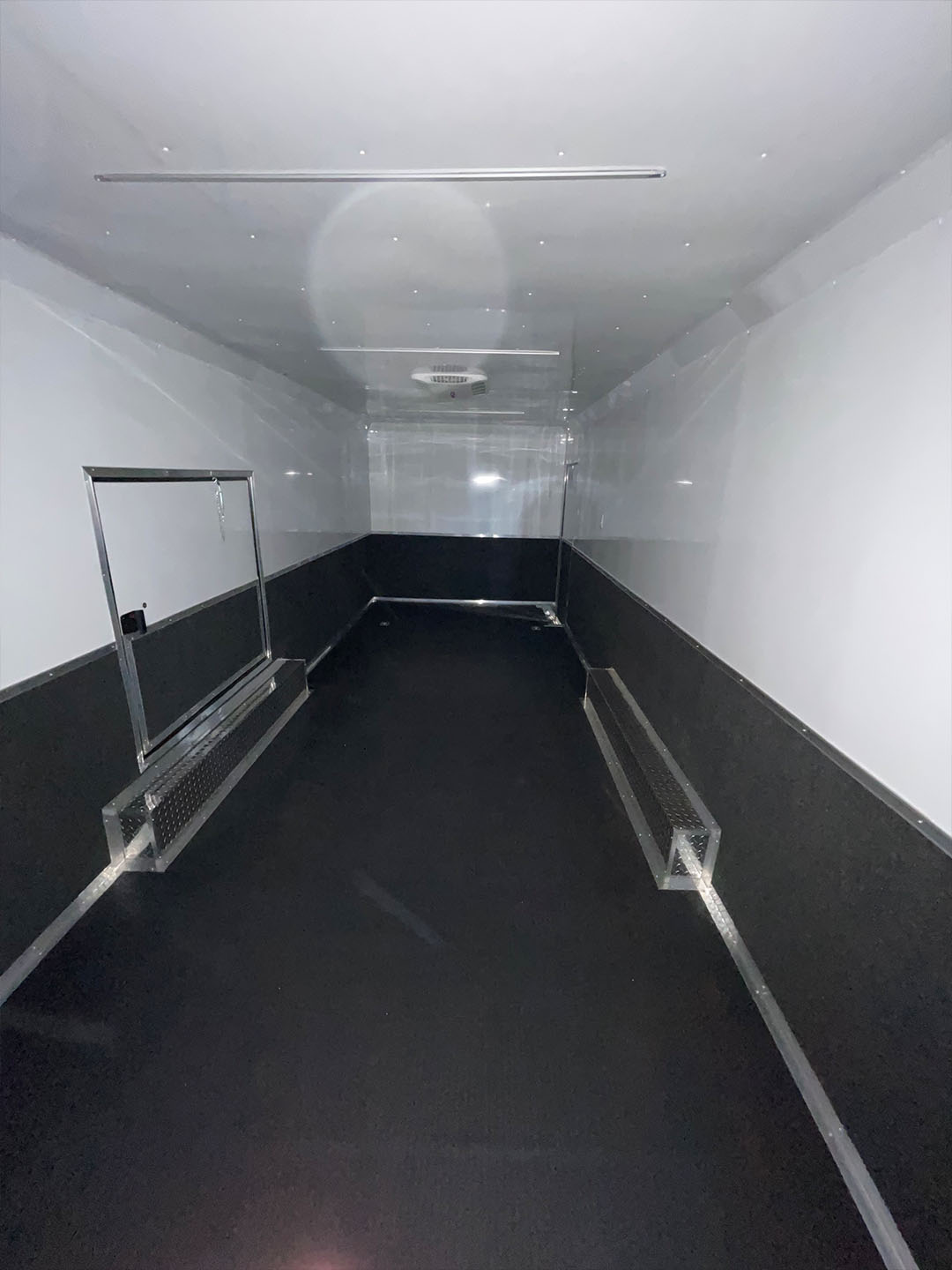 The image shows an interior view of a narrow corridor with white walls and a reflective floor, featuring a metal railing on one side and a black carpeted area on the other.