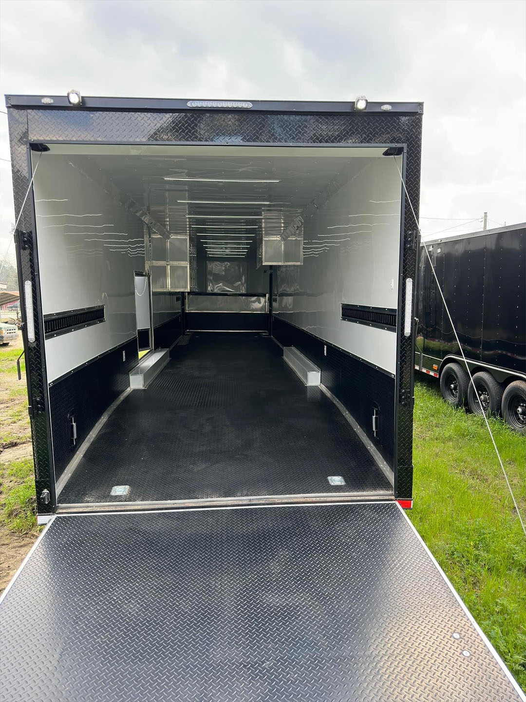 The image shows a large black enclosed trailer parked on grass with its door open, revealing an interior space that appears to be empty.