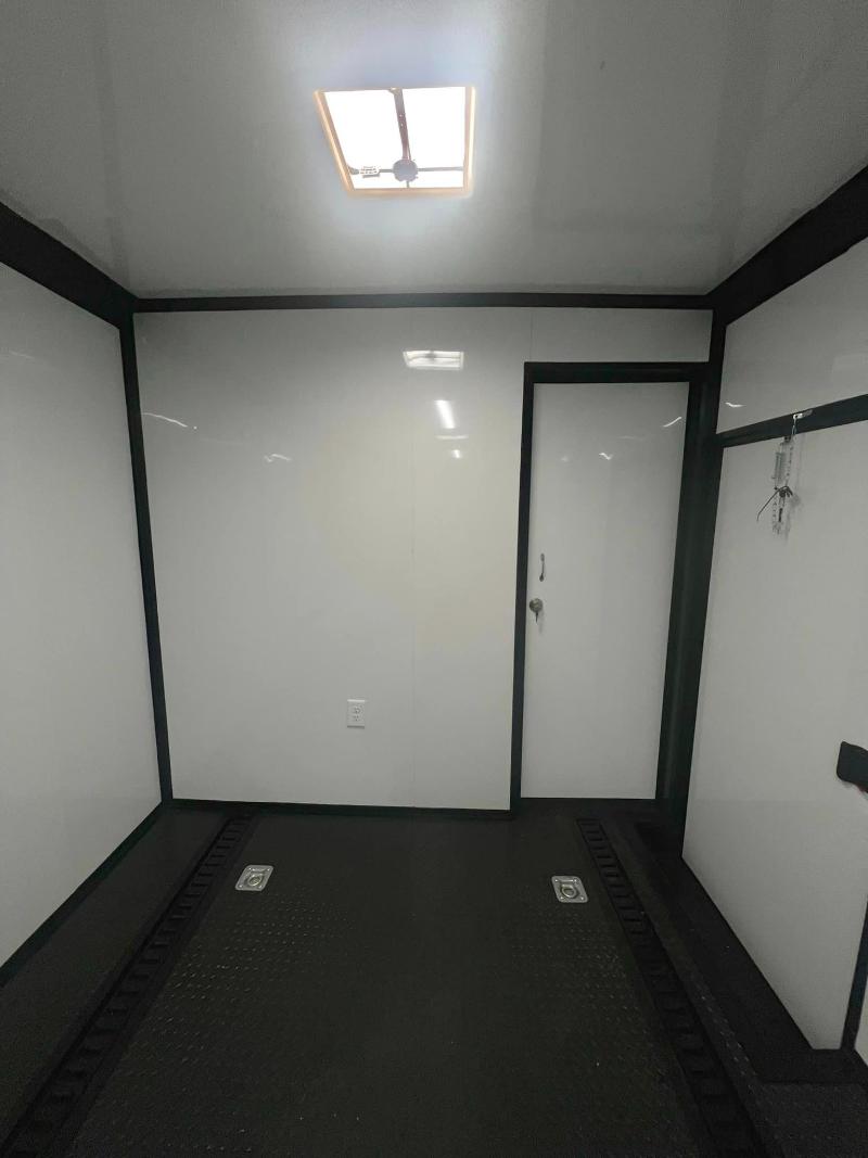 The image shows an interior view of a clean, empty room with white walls, a high ceiling, and a single light fixture on the ceiling. There is a white door at one end of the room, and a small window near the top left corner. The floor appears to be a dark color, possibly black or gray.