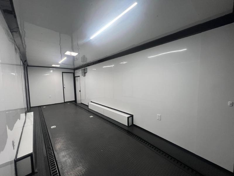 This image shows an interior view of a large, empty room with white walls, a black floor, and various pieces of equipment on the right side, including what appears to be a vehicle chassis and mechanical components, suggesting this space may be used for automotive manufacturing or assembly.
