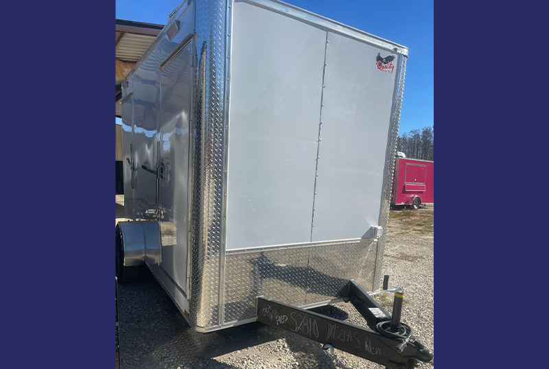 The image shows a silver-colored enclosed trailer parked on a gravel surface, with a clear sky in the background.