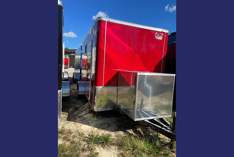 The image shows a red trailer parked on a gravel surface with a clear sky background.