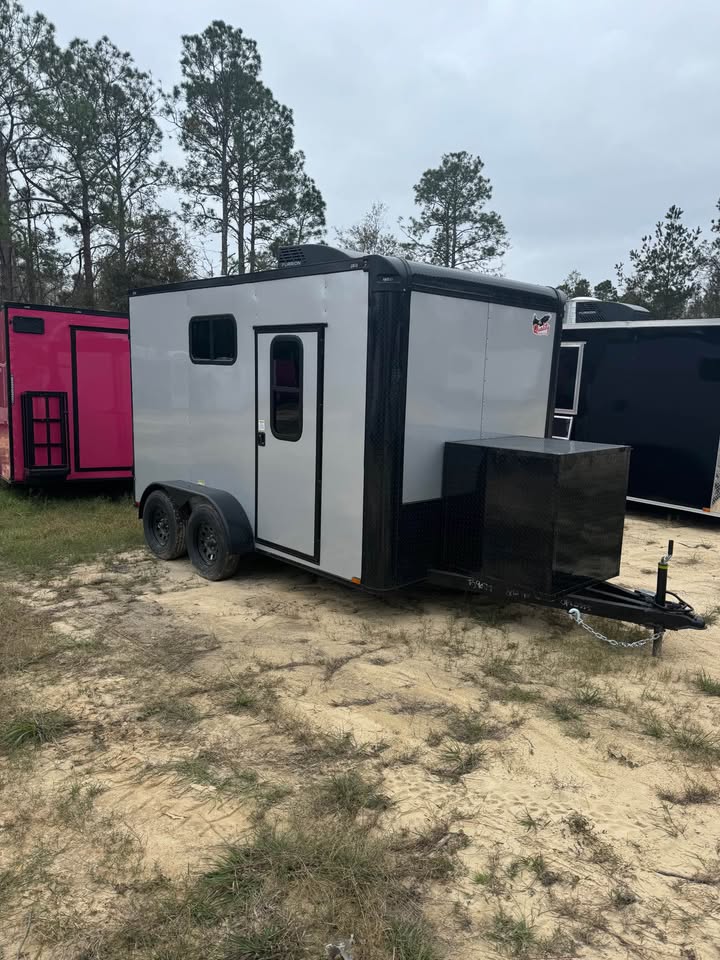 The image shows a large white horse trailer parked on a dirt lot with grassy areas and trees in the background.