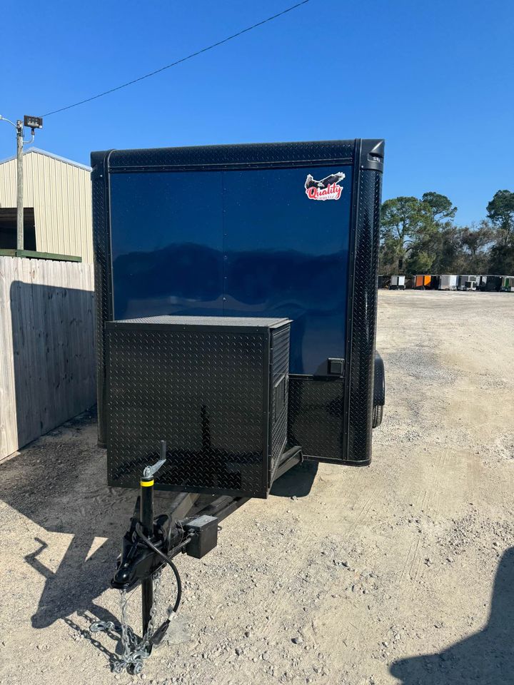 The image shows a black trailer parked on a gravel surface with a clear sky in the background.