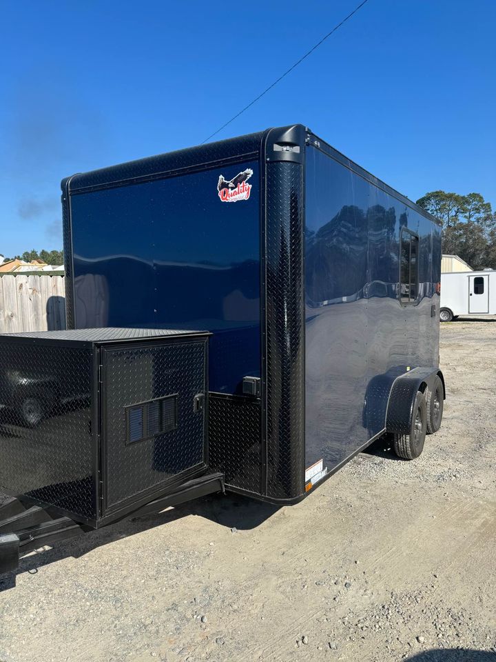 The image depicts a large black enclosed trailer parked on a gravel surface with a clear blue sky in the background.