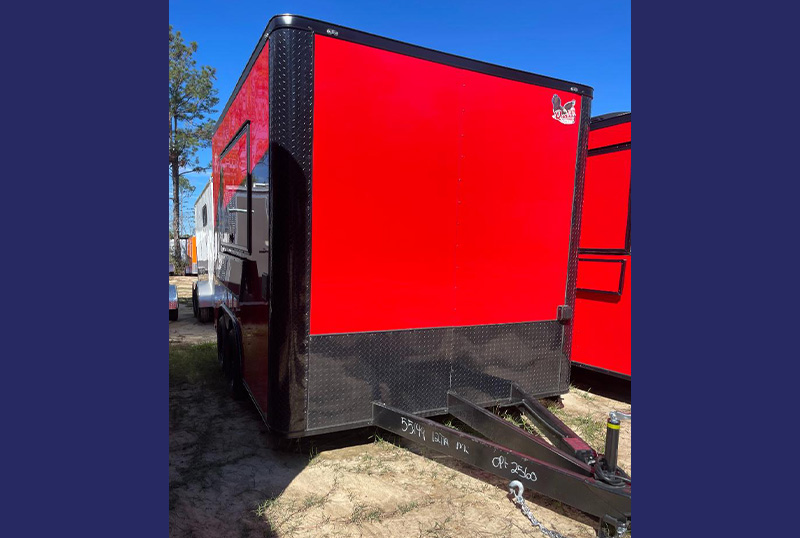 The image shows a red trailer parked on a dirt surface with a clear blue sky in the background.