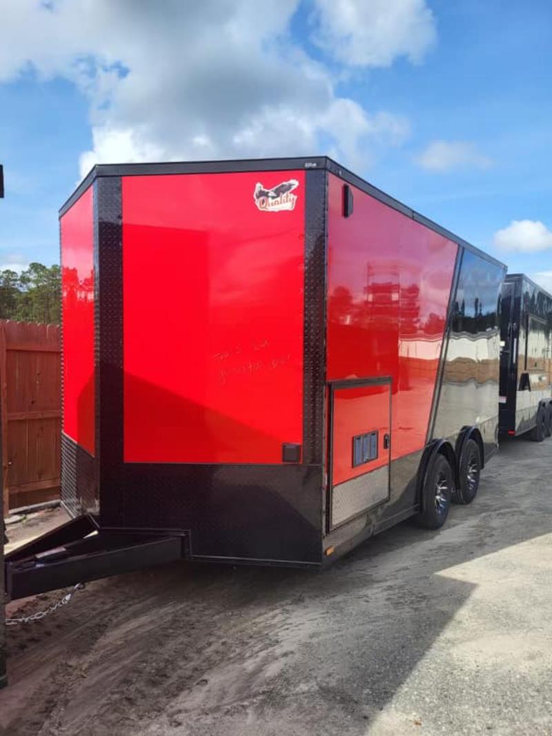 The image shows a large red and black enclosed trailer parked on a gravel surface with a clear sky in the background.