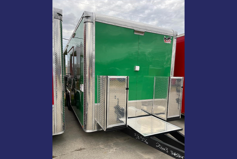 The image shows a large, green enclosed trailer with a silver toolbox on the back, parked outdoors during daylight hours.