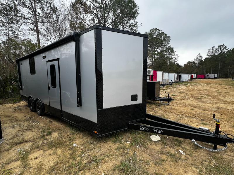 The image shows a large, gray, enclosed trailer parked on a dirt surface with trees in the background.