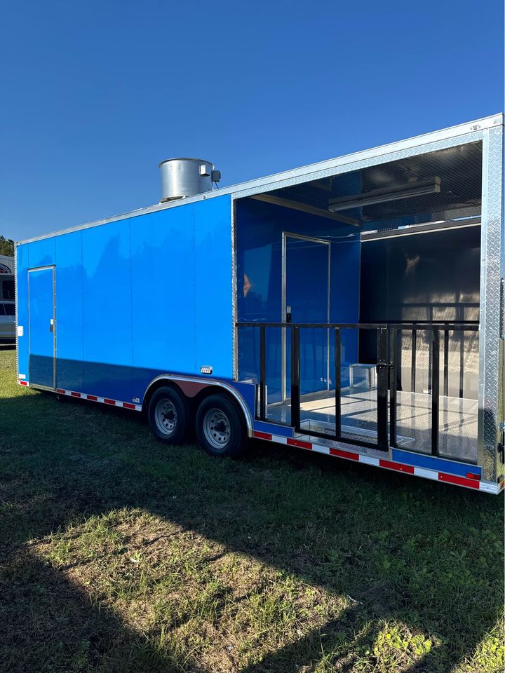 The image shows a large blue food truck parked on a grassy field with clear skies above.