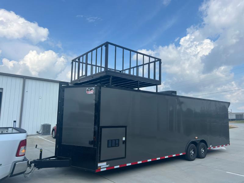 This is a photograph of a large black enclosed trailer being towed by a white pickup truck, with a black metal platform on top featuring railings and a staircase leading up to it.