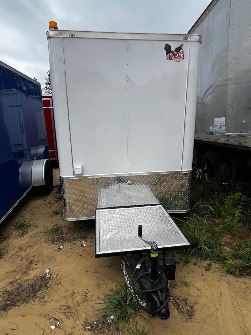 The image shows a white utility trailer parked on a dirt surface with a blue truck visible behind it.