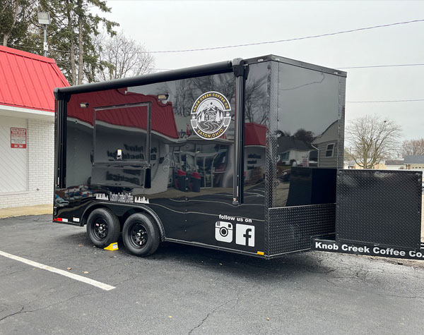 The image shows a black food truck parked in a lot with a clear sky background.