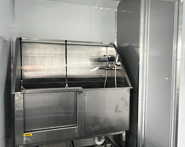 The image shows a stainless steel industrial kitchen appliance with a large metal door and a control panel on the right side, situated inside a clean room environment with white walls and a gray floor.