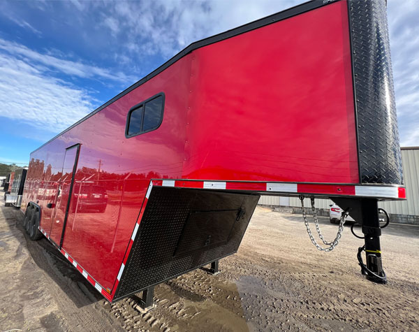 The image shows a large red enclosed trailer parked on a gravel surface with a clear sky in the background.