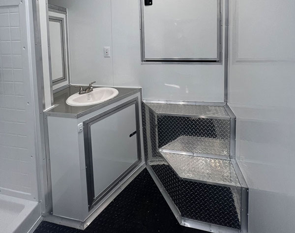 The image shows a compact bathroom within a mobile trailer or camper, featuring a sink, mirror, and storage area with a metal door.