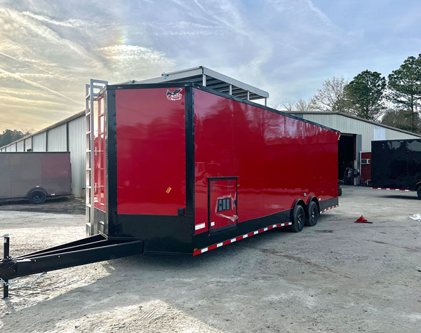 The image shows a large red semi-trailer truck parked on a concrete surface with a building and trees in the background.