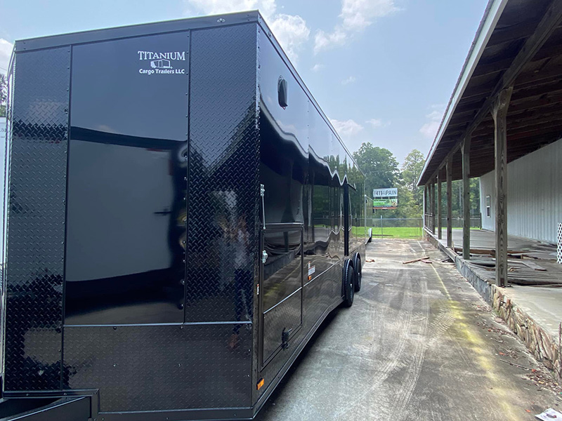 The image shows a black enclosed trailer parked on a paved lot next to a building with a visible sign.