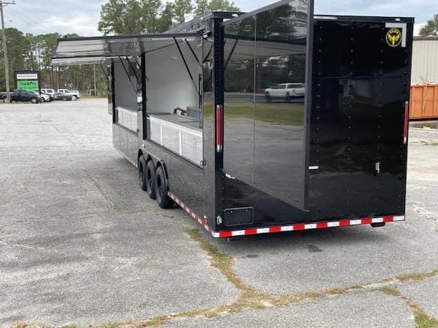 The image features a black food truck parked on an asphalt surface with a clear sky above.