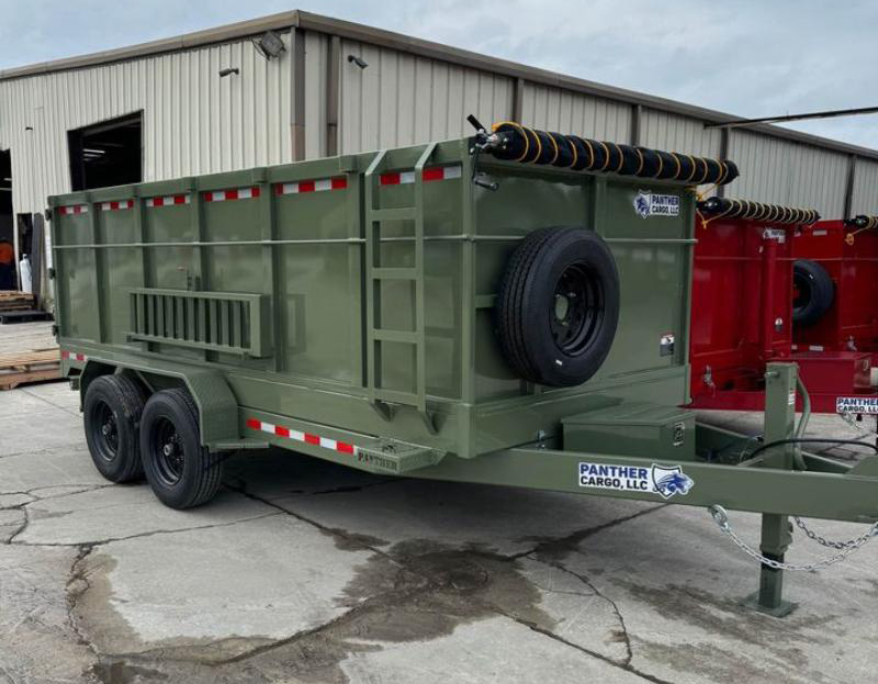 This image shows a large military-style green utility truck parked on an asphalt surface next to a building with a corrugated metal roof, featuring a trailer attached to its rear.